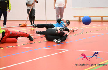 Show 2 goalball players defending the goal on a goalball court with the blue ball just being defelected off an defender and its played in a sports hall
