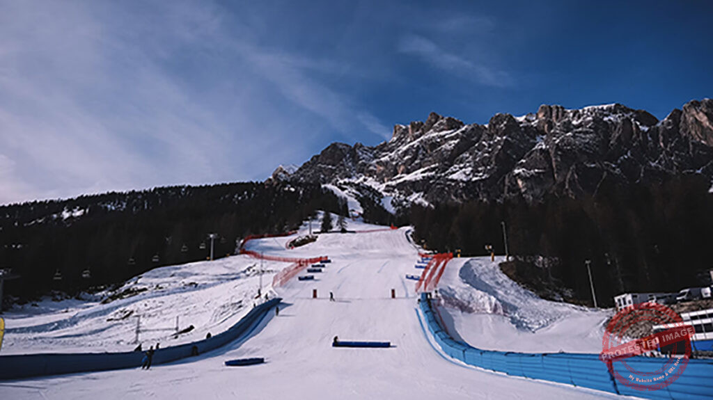 This photo is of Milano Cortina in preperation for the Paralympics in 100 days 6th March. Shows a alrge mountain range with a ski slop in the middle with blue barriers eather side on a sunny day